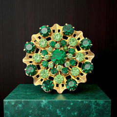 Vintage 1950s gold tone brooch with a ruffled sunburst design, featuring concentric rings of emerald and peridot rhinestones and a large green center stone; bold mid-century costume jewelry with layered sparkle and sculptural detail.