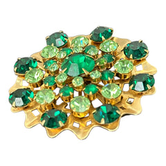 Vintage 1950s gold tone brooch with a ruffled sunburst design, featuring concentric rings of emerald and peridot rhinestones and a large green center stone; bold mid-century costume jewelry with layered sparkle and sculptural detail.