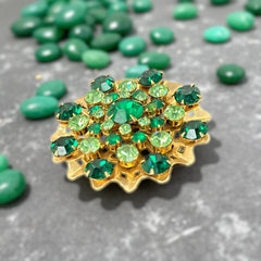 Vintage 1950s gold tone brooch with a ruffled sunburst design, featuring concentric rings of emerald and peridot rhinestones and a large green center stone; bold mid-century costume jewelry with layered sparkle and sculptural detail.