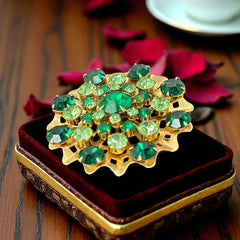 Vintage 1950s gold tone brooch with a ruffled sunburst design, featuring concentric rings of emerald and peridot rhinestones and a large green center stone; bold mid-century costume jewelry with layered sparkle and sculptural detail.