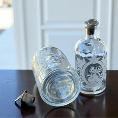 Set of two vintage etched glass apothecary bottles with frosted floral designs and cork stoppers topped with metal rings, photographed in natural light.