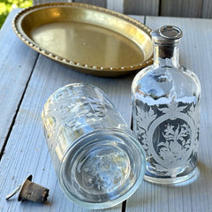 Set of two vintage etched glass apothecary bottles with frosted floral designs and cork stoppers topped with metal rings, photographed in natural light.