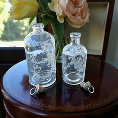 Set of two vintage etched glass apothecary bottles with frosted floral designs and cork stoppers topped with metal rings, photographed in natural light.