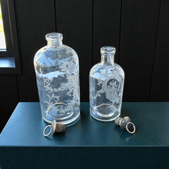 Set of two vintage etched glass apothecary bottles with frosted floral designs and cork stoppers topped with metal rings, photographed in natural light.