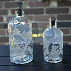 Set of two vintage etched glass apothecary bottles with frosted floral designs and cork stoppers topped with metal rings, photographed in natural light.