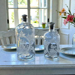 Set of two vintage etched glass apothecary bottles with frosted floral designs and cork stoppers topped with metal rings, photographed in natural light.