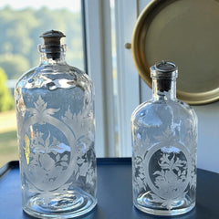 Set of two vintage etched glass apothecary bottles with frosted floral designs and cork stoppers topped with metal rings, photographed in natural light.