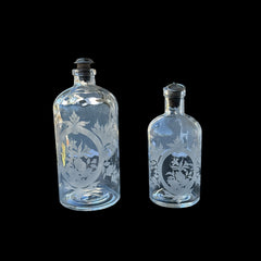 Set of two vintage etched glass apothecary bottles with frosted floral designs and cork stoppers topped with metal rings, photographed in natural light.