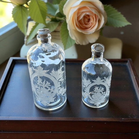 Set of two vintage etched glass apothecary bottles with frosted floral designs and cork stoppers topped with metal rings, photographed in natural light.