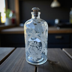 Set of two vintage etched glass apothecary bottles with frosted floral designs and cork stoppers topped with metal rings, photographed in natural light.