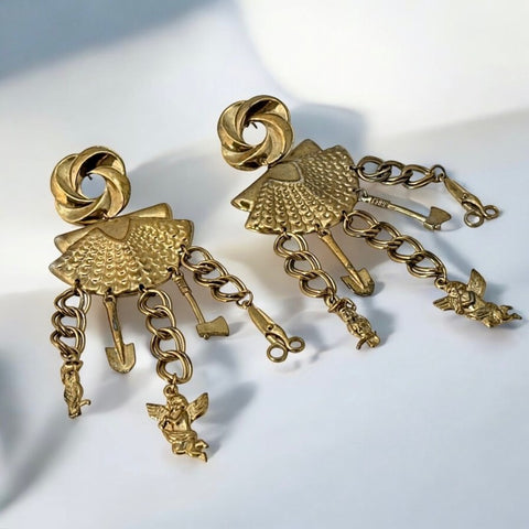 Vintage 1980s French stamped brass dangle earrings featuring a scallop shell centerpiece with eclectic dangling charms, including an owl, shovel, putti angel, axe, and scissors. Unique statement jewelry with a bold boho style, rich patina, and artistic character.