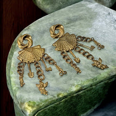 Vintage 1980s French stamped brass dangle earrings featuring a scallop shell centerpiece with eclectic dangling charms, including an owl, shovel, putti angel, axe, and scissors. Unique statement jewelry with a bold boho style, rich patina, and artistic character.