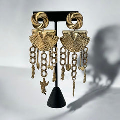 Vintage 1980s French stamped brass dangle earrings featuring a scallop shell centerpiece with eclectic dangling charms, including an owl, shovel, putti angel, axe, and scissors. Unique statement jewelry with a bold boho style, rich patina, and artistic character.