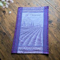 French jacquard cotton tea towel in lavender, violet, and white featuring Provençal lavender fields, farmhouse, and “La Provence” lettering. Made in France.