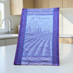 French jacquard cotton tea towel in lavender, violet, and white featuring Provençal lavender fields, farmhouse, and “La Provence” lettering. Made in France.