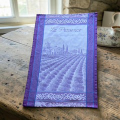 French jacquard cotton tea towel in lavender, violet, and white featuring Provençal lavender fields, farmhouse, and “La Provence” lettering. Made in France.