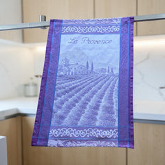 French jacquard cotton tea towel in lavender, violet, and white featuring Provençal lavender fields, farmhouse, and “La Provence” lettering. Made in France.