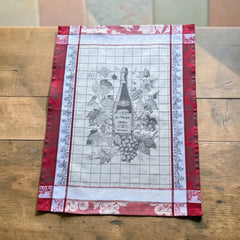 French jacquard cotton tea towel in red, white, and soft gray beige featuring a Le Vieux Chêne wine bottle surrounded by grapes and vine leaves.
