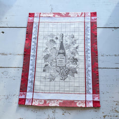 French jacquard cotton tea towel in red, white, and soft gray beige featuring a Le Vieux Chêne wine bottle surrounded by grapes and vine leaves.