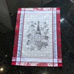 French jacquard cotton tea towel in red, white, and soft gray beige featuring a Le Vieux Chêne wine bottle surrounded by grapes and vine leaves.