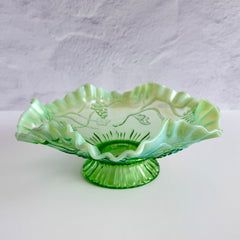 Antique green opalescent glass compote bowl with a ruffled rim, grape and leaf pattern, and a round pedestal foot, photographed against a light background.