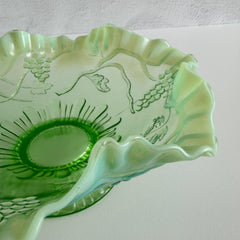 Antique green opalescent glass compote bowl with a ruffled rim, grape and leaf pattern, and a round pedestal foot, photographed against a light background.