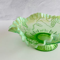 Antique green opalescent glass compote bowl with a ruffled rim, grape and leaf pattern, and a round pedestal foot, photographed against a light background.