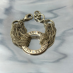 Gold tone Guess bracelet with multi-strand chains and a central ring engraved with ‘GUESS’ and accented by clear rhinestones, shown on neutral backdrop.