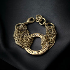Gold tone Guess bracelet with multi-strand chains and a central ring engraved with ‘GUESS’ and accented by clear rhinestones, shown on neutral backdrop.