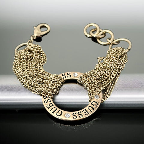 Gold tone Guess bracelet with multi-strand chains and a central ring engraved with ‘GUESS’ and accented by clear rhinestones, shown on neutral backdrop.
