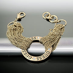Gold tone Guess bracelet with multi-strand chains and a central ring engraved with ‘GUESS’ and accented by clear rhinestones, shown on neutral backdrop.