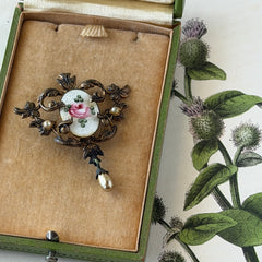 Vintage white guilloché enamel brooch with pink rose motif, faux pearls, and ornate antiqued gold tone filigree frame with teardrop pearl accent.