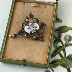 Vintage white guilloché enamel brooch with pink rose motif, faux pearls, and ornate antiqued gold tone filigree frame with teardrop pearl accent.