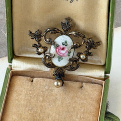 Vintage white guilloché enamel brooch with pink rose motif, faux pearls, and ornate antiqued gold tone filigree frame with teardrop pearl accent.