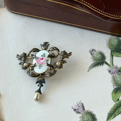 Vintage white guilloché enamel brooch with pink rose motif, faux pearls, and ornate antiqued gold tone filigree frame with teardrop pearl accent.