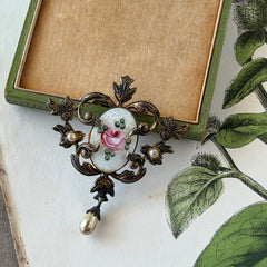 Vintage white guilloché enamel brooch with pink rose motif, faux pearls, and ornate antiqued gold tone filigree frame with teardrop pearl accent.