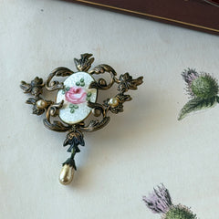 Vintage white guilloché enamel brooch with pink rose motif, faux pearls, and ornate antiqued gold tone filigree frame with teardrop pearl accent.