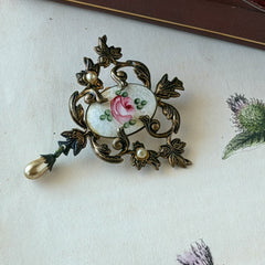 Vintage white guilloché enamel brooch with pink rose motif, faux pearls, and ornate antiqued gold tone filigree frame with teardrop pearl accent.