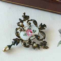 Vintage white guilloché enamel brooch with pink rose motif, faux pearls, and ornate antiqued gold tone filigree frame with teardrop pearl accent.