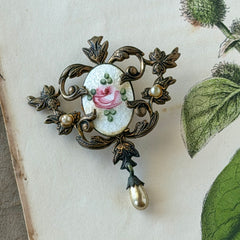 Vintage white guilloché enamel brooch with pink rose motif, faux pearls, and ornate antiqued gold tone filigree frame with teardrop pearl accent.