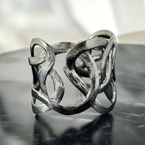 Textured dark silver tone cuff bracelet with interwoven, ribbon-like bands forming a sculptural openwork design; hammered finish adds depth and dimension to the modern, unsigned piece.