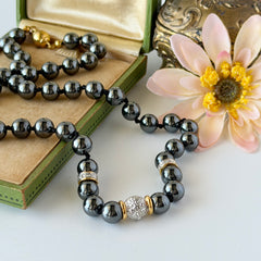 Vintage hematite bead necklace with gold‑tone and silver‑tone accents, hand‑knotted with slide ball clasp, displayed on green box.