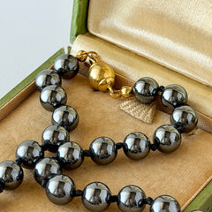 Vintage hematite bead necklace with gold‑tone and silver‑tone accents, hand‑knotted with slide ball clasp, displayed on green box.