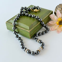 Vintage hematite bead necklace with gold‑tone and silver‑tone accents, hand‑knotted with slide ball clasp, displayed on green box.