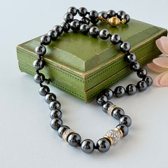 Vintage hematite bead necklace with gold‑tone and silver‑tone accents, hand‑knotted with slide ball clasp, displayed on green box.