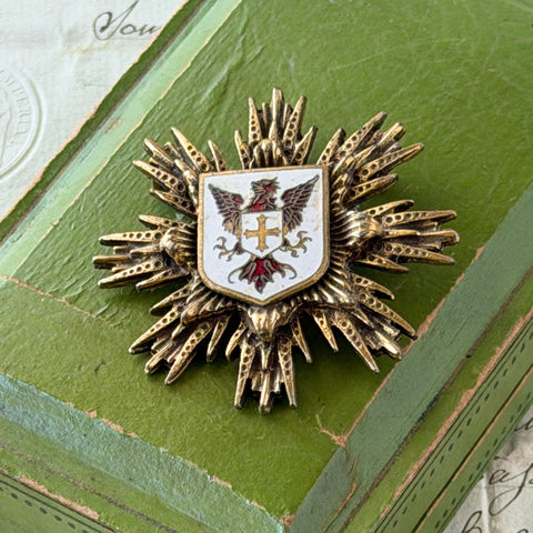 Vintage gold‑tone heraldic brooch with white enamel shield and red‑gold griffin coat of arms, photographed on antique paper and boxes.