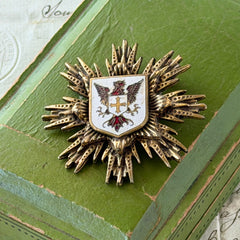 Vintage gold‑tone heraldic brooch with white enamel shield and red‑gold griffin coat of arms, photographed on antique paper and boxes.