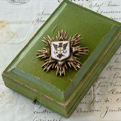 Vintage gold‑tone heraldic brooch with white enamel shield and red‑gold griffin coat of arms, photographed on antique paper and boxes.