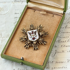 Vintage gold‑tone heraldic brooch with white enamel shield and red‑gold griffin coat of arms, photographed on antique paper and boxes.