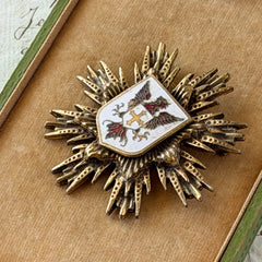 Vintage gold‑tone heraldic brooch with white enamel shield and red‑gold griffin coat of arms, photographed on antique paper and boxes.